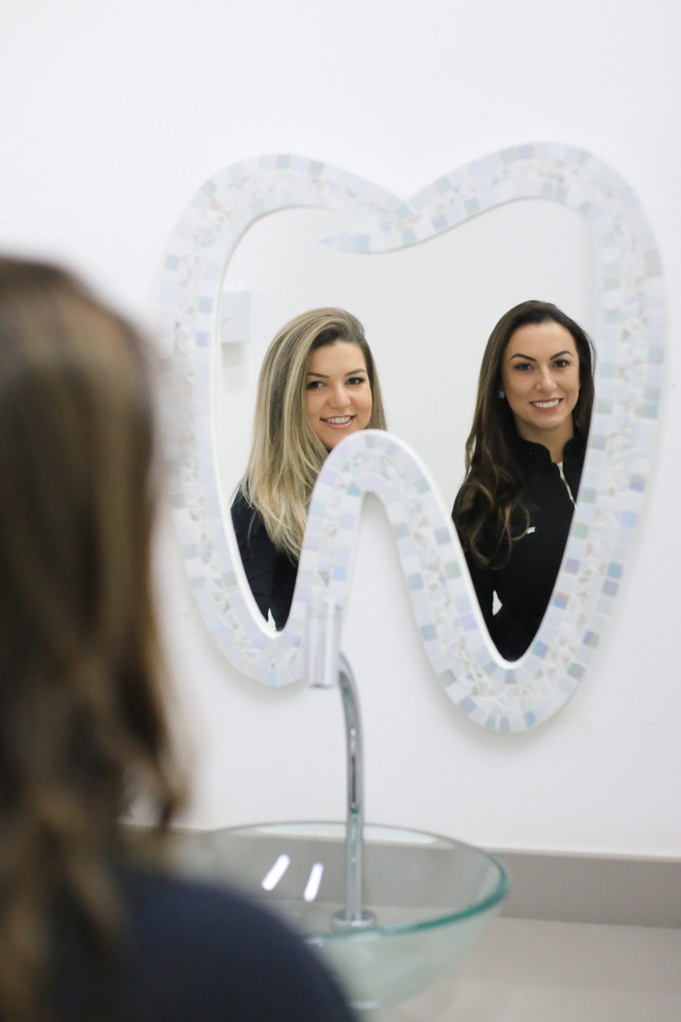 Espelho em forma de dente refletindo duas dentistas sorrindo